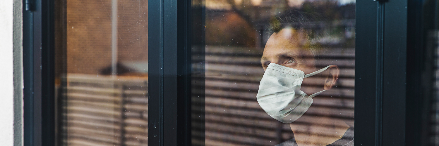 man peering out window wearing surgical mask
