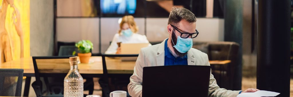 two professionals sitting at desks at public workspace wearing surgical masks while working on laptops.