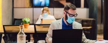 two professionals sitting at desks at public workspace wearing surgical masks while working on laptops.