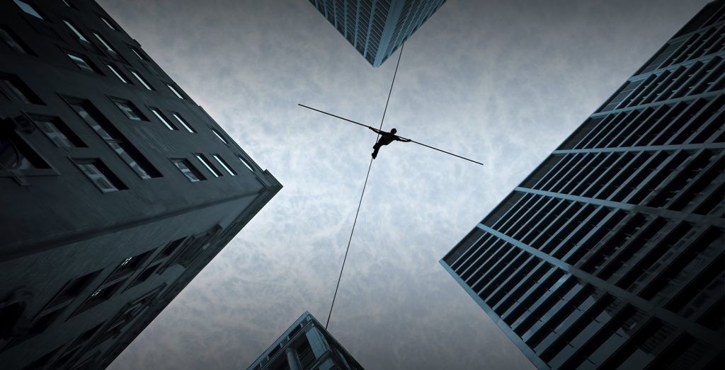 ground view of man tightrope walking across the roofs of two high rise buildings